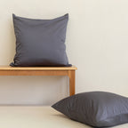 Two grey European pillowcases against a neutral background. One on a timber bench, one on the floor.