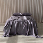 Bed with grey bedding and pillows against a beige curtain background.