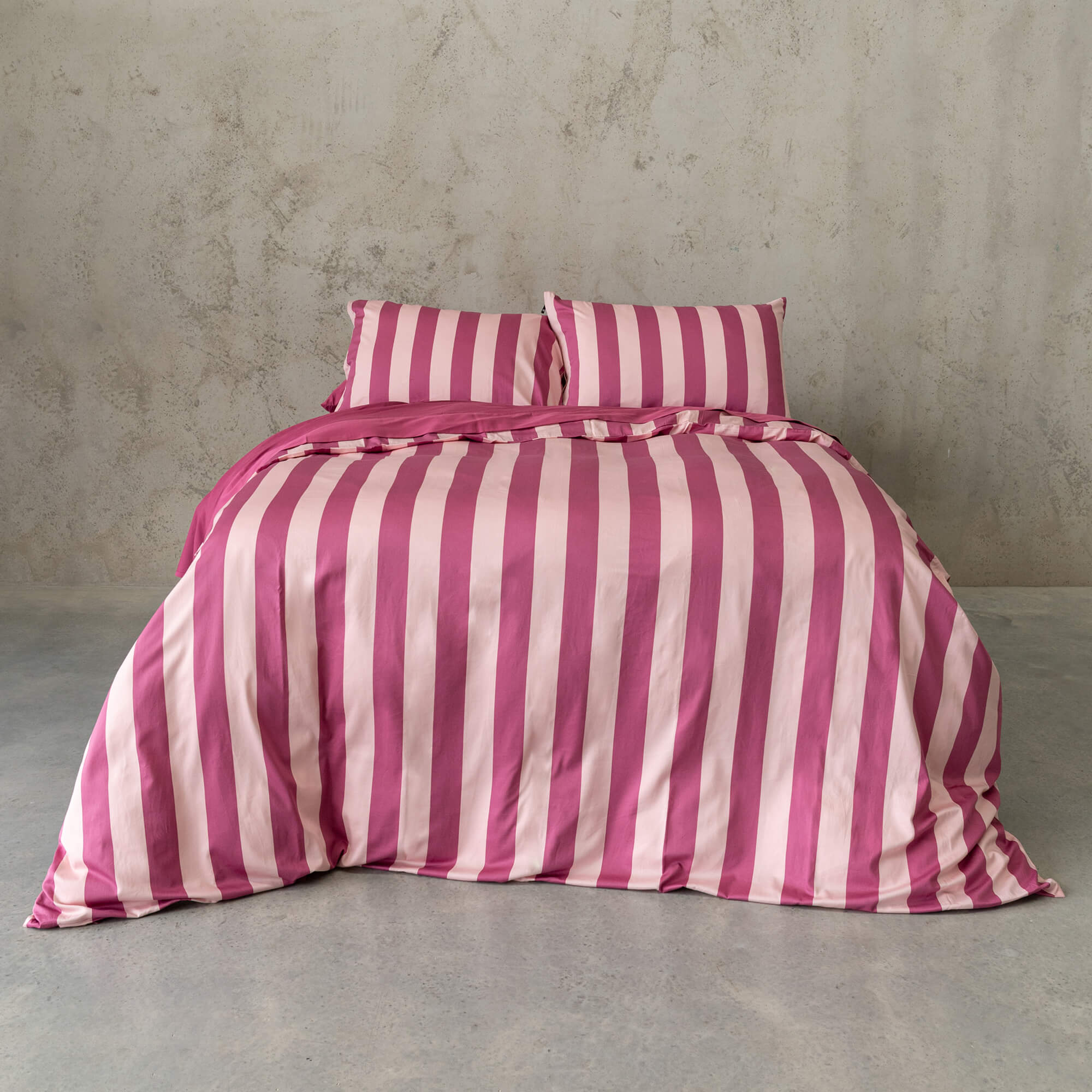 Raspberry pink stripe quilt cover set on a bed, against a neutral concrete look backdrop. 