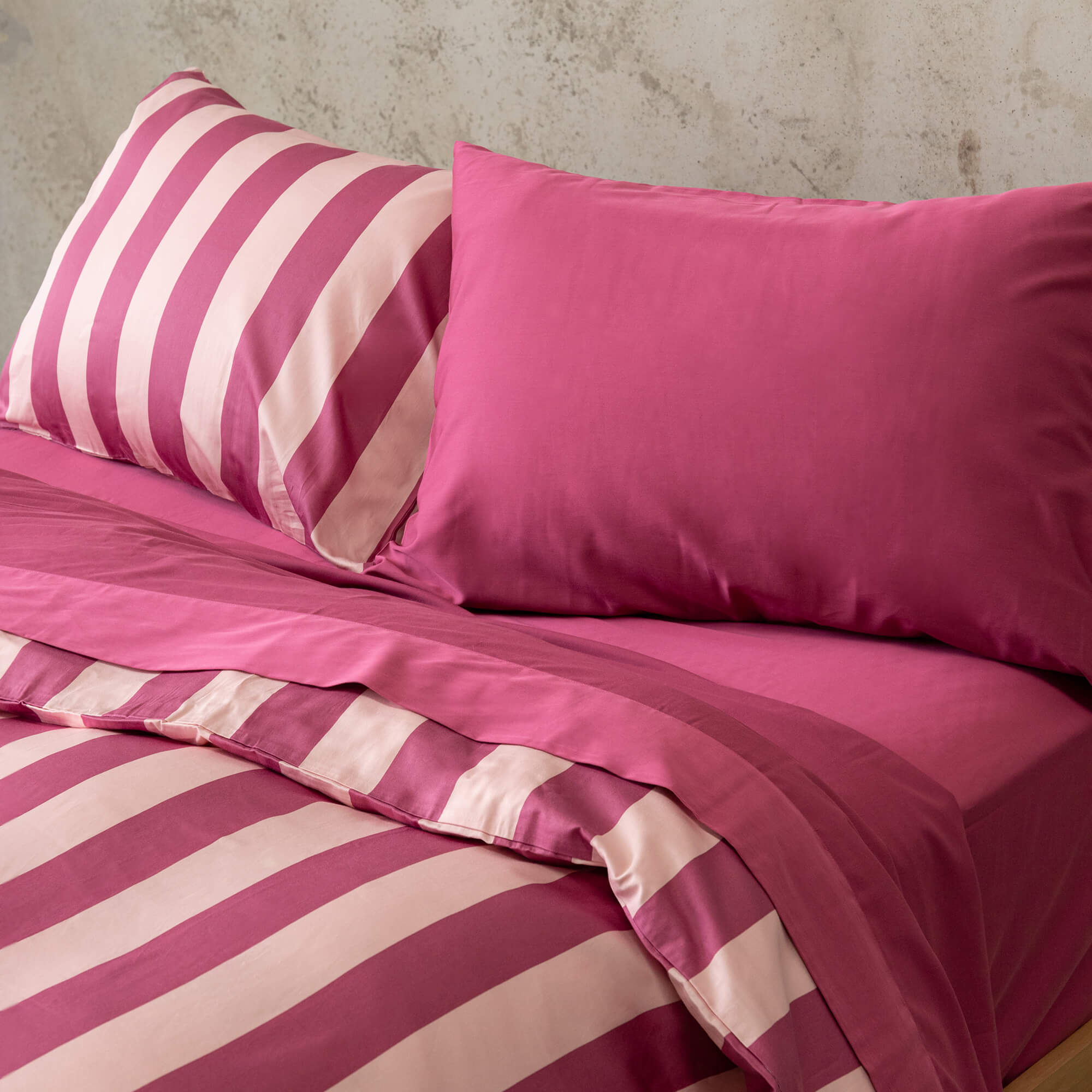 Raspberry pink stripe quilt cover set on a bed, against a neutral concrete look backdrop. 
