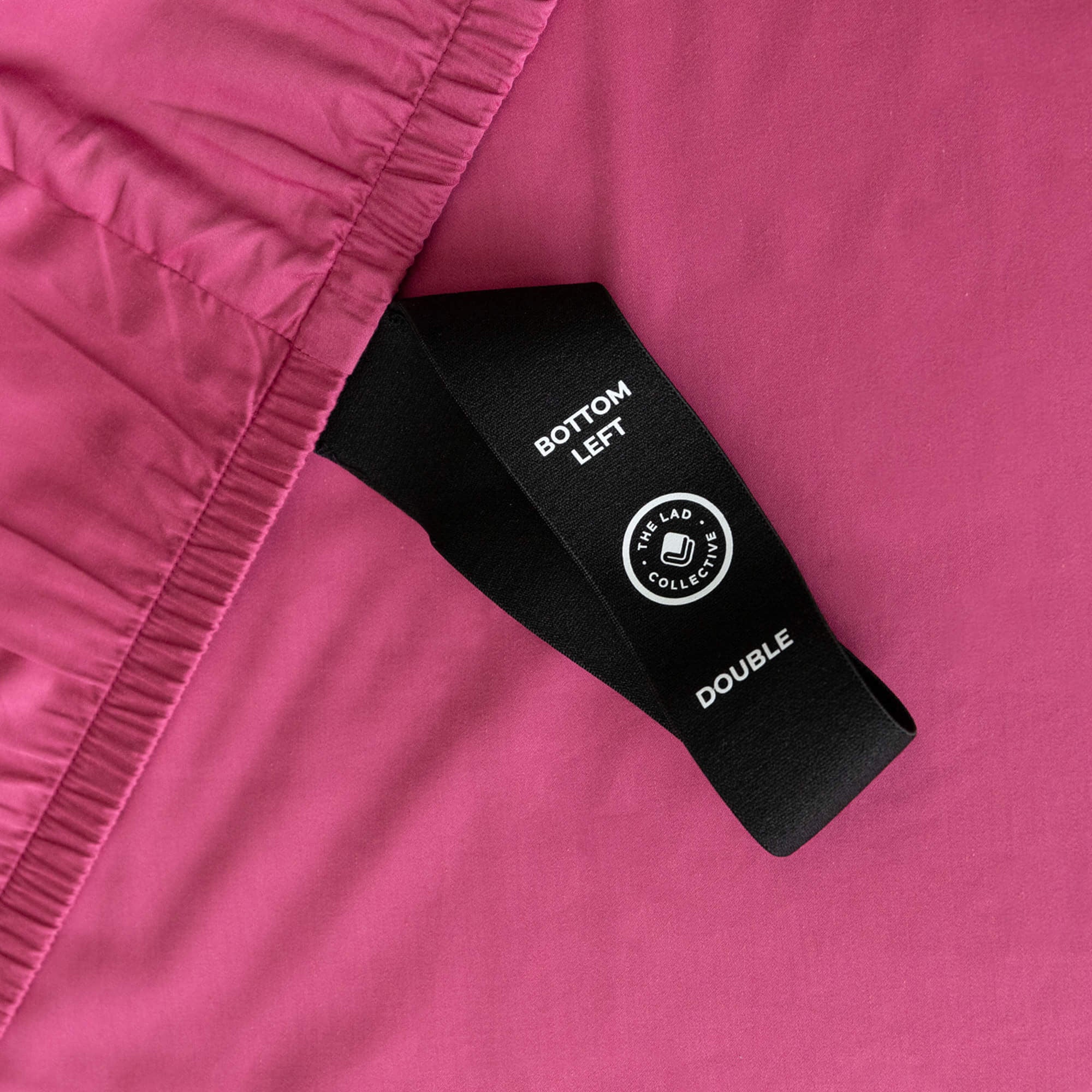 Raspberry pink fitted sheet labelled corner loop strap that reads 'Bottom Left' and 'Double'.