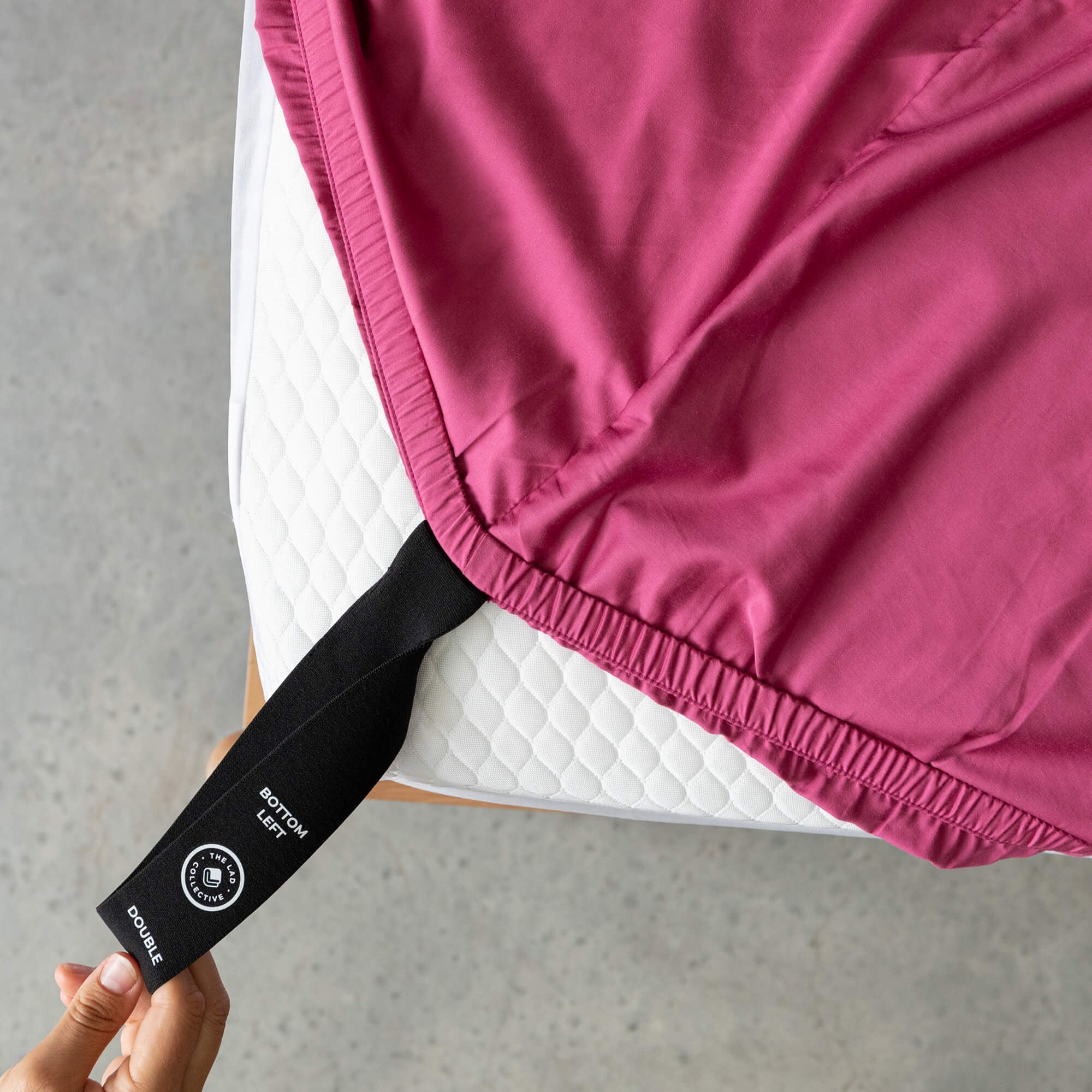 Raspberry pink fitted sheet labelled corner loop strap that reads 'Bottom Left' and 'Double', being pulled onto a mattress corner by a hand.