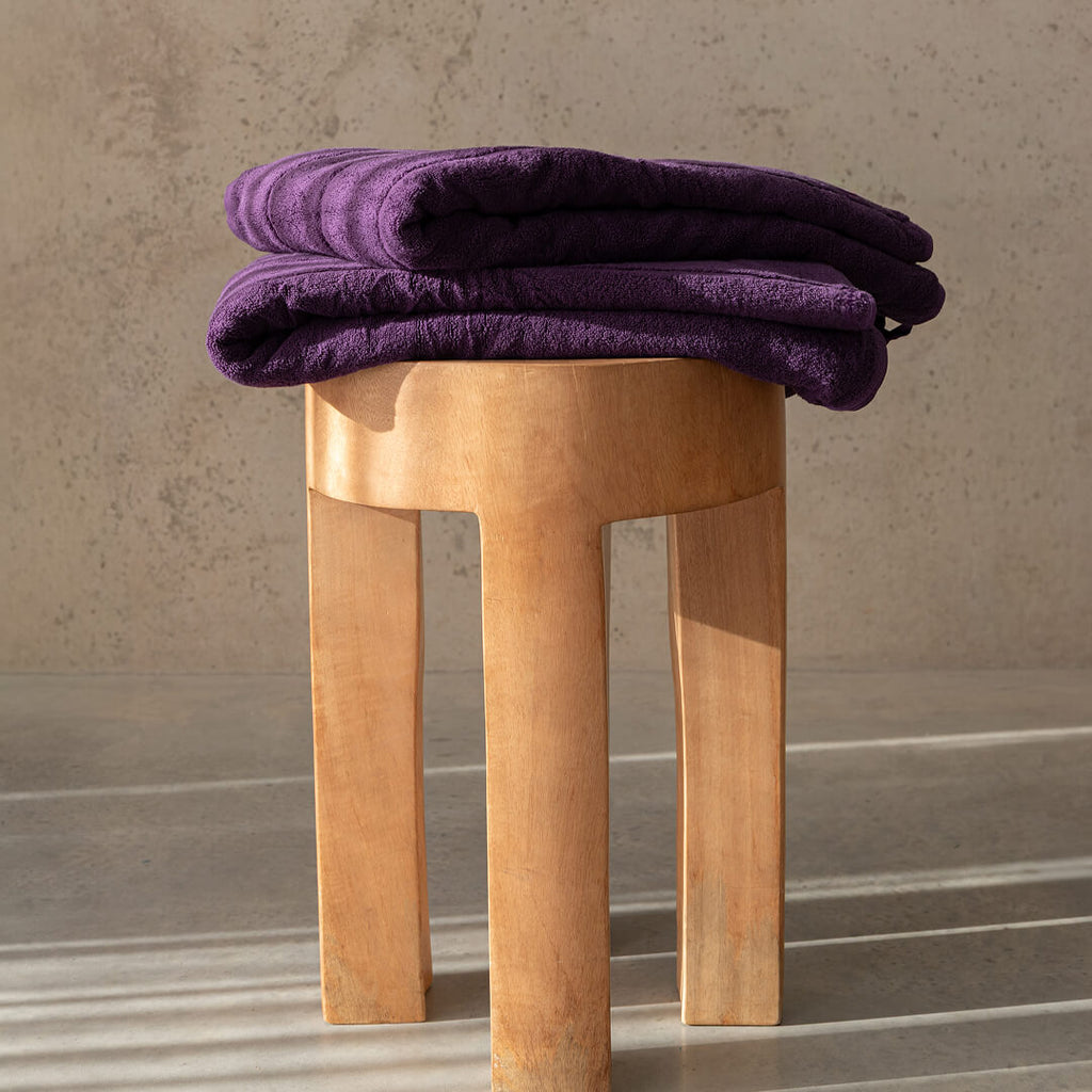 Purple bath towels folded on a wooden stool against a neutral background.