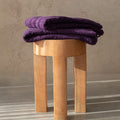 Purple bath towels folded on a wooden stool against a neutral background.