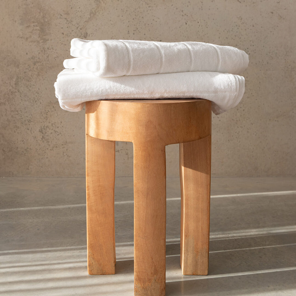 Two white bath towels folded on top of a timber stool, with a concrete background.
