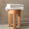 Two white bath towels folded on top of a timber stool, with a concrete background.