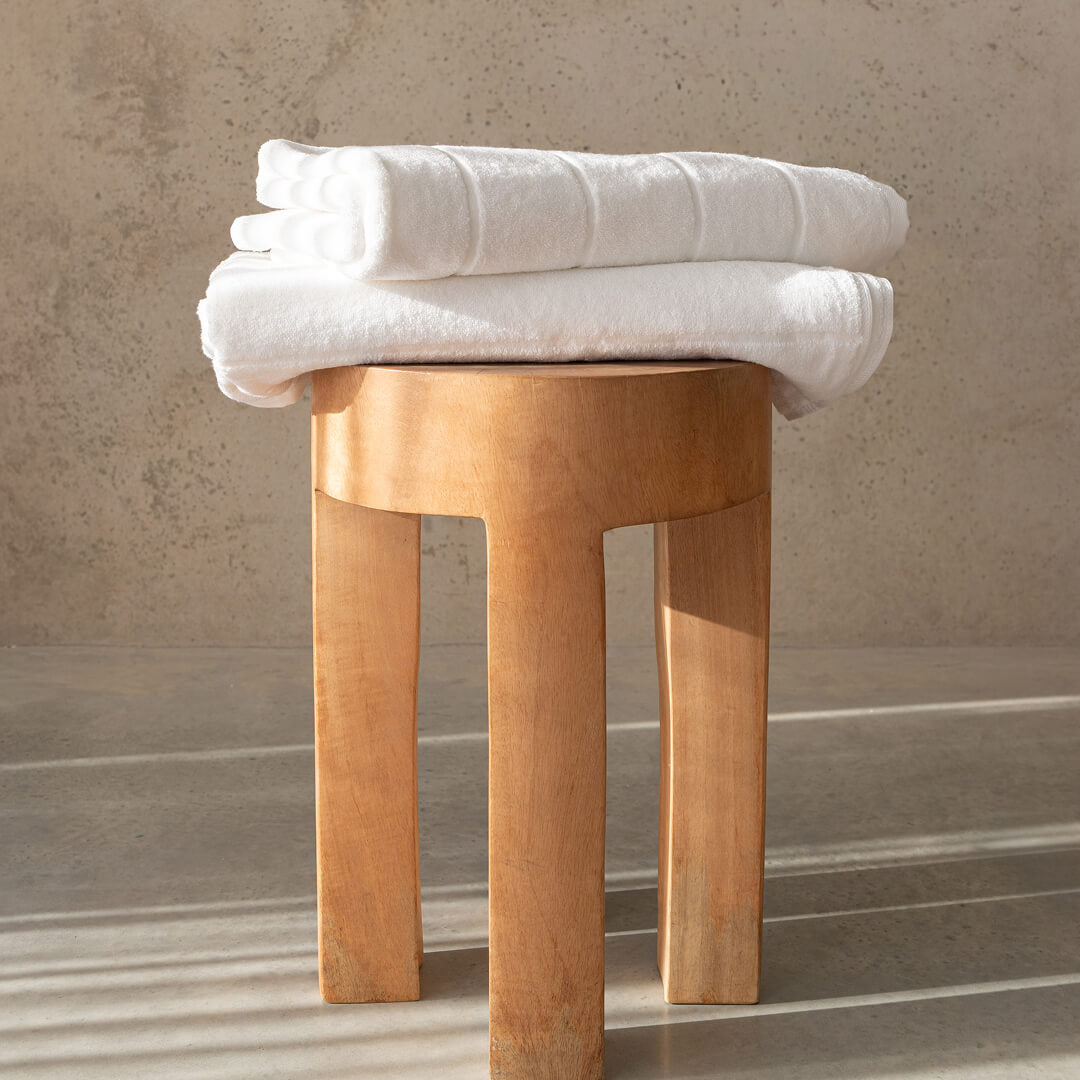 Two white bath towels folded on top of a timber stool, with a concrete background.
