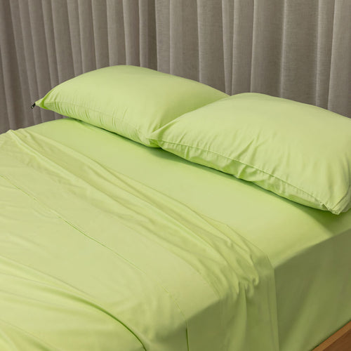Green bed sheets and pillows on a bed with a neutral curtain background.