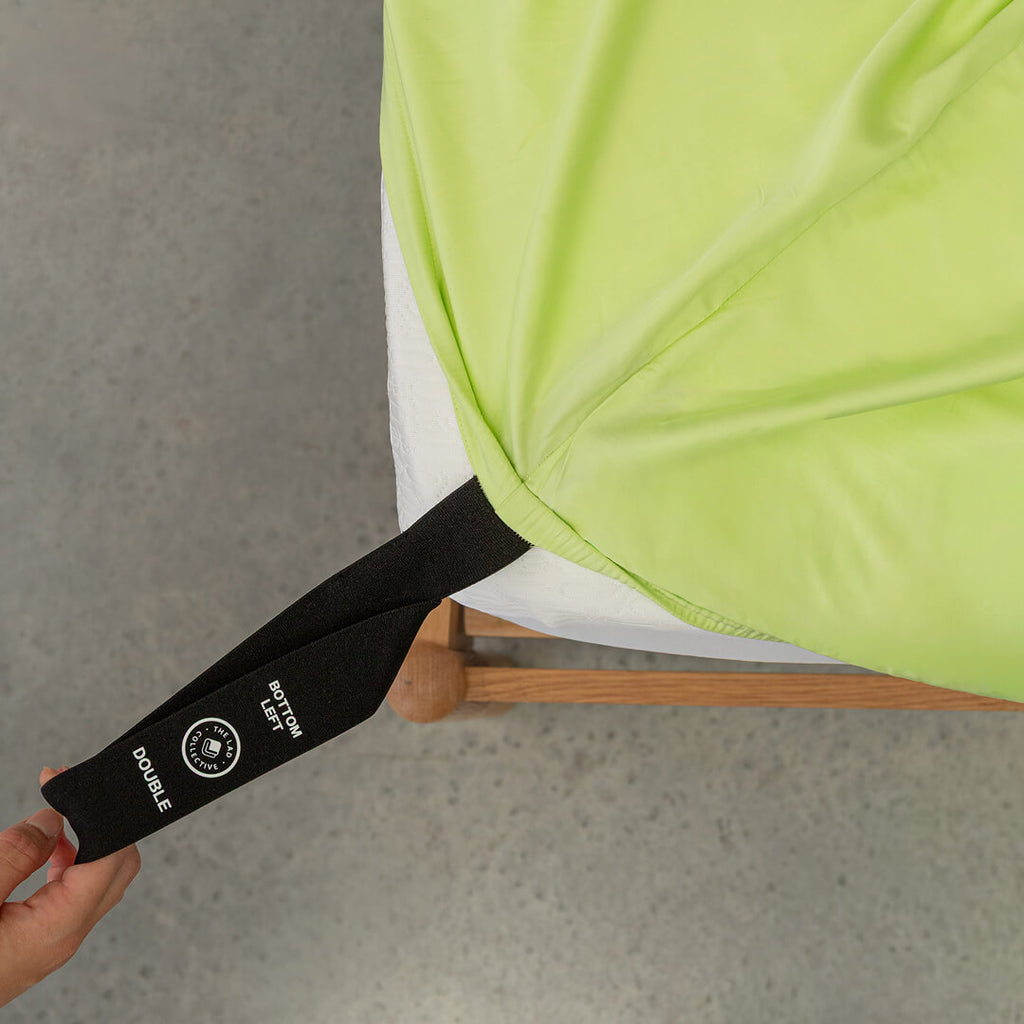 Green fitted sheet with a black strap and visible brand logo being pulled by a hand onto the mattress.