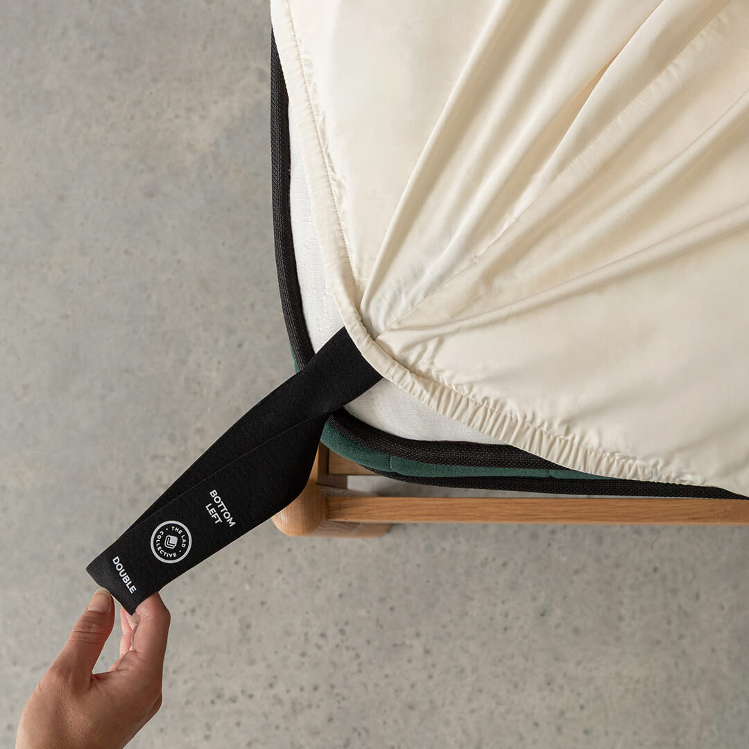 Hand pulling the black labelled corner loop strap that says 'Bottom Left' on cream fitted sheet.