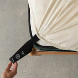 Hand pulling the black labelled corner loop strap that says 'Bottom Left' on cream fitted sheet.