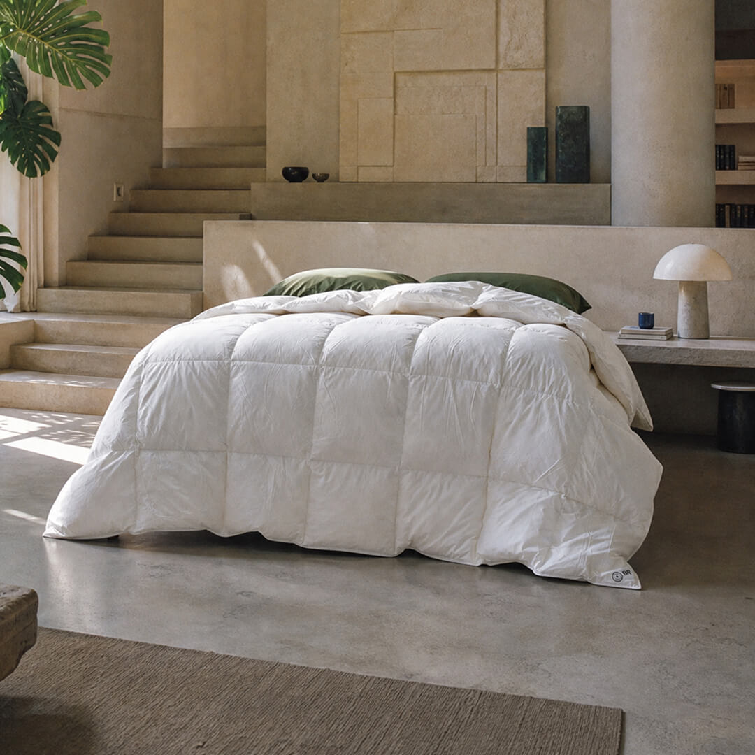 White comforter on a bed in a modern bedroom with concrete floor and stone walls.