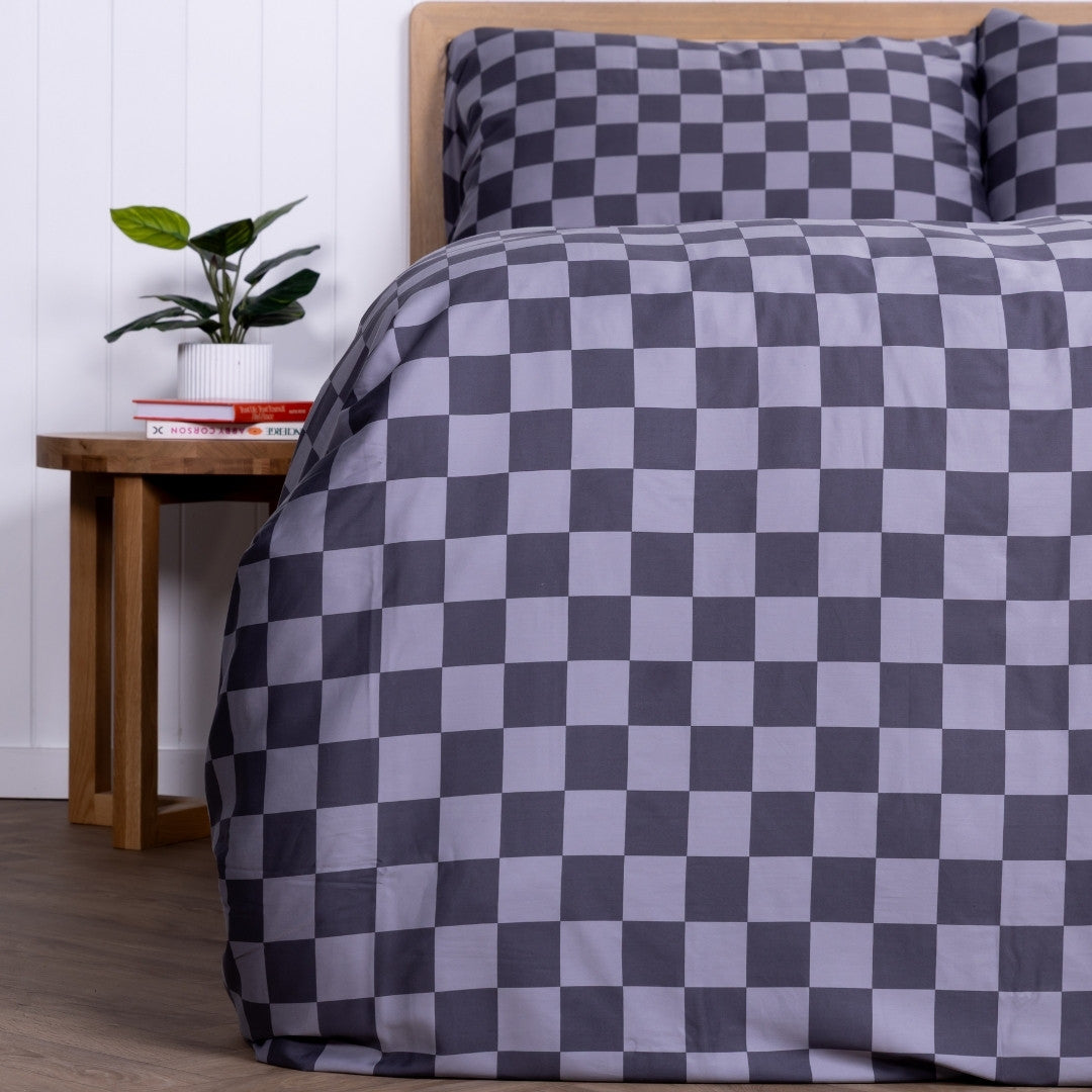 Grey checkered bedspread on a bed with a wooden side table and plant in the background.