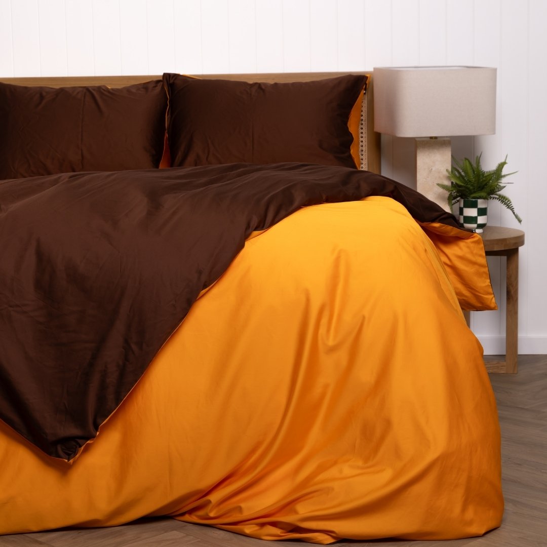 Brown and orange bedding set on a bed with a lamp and plant in the background.